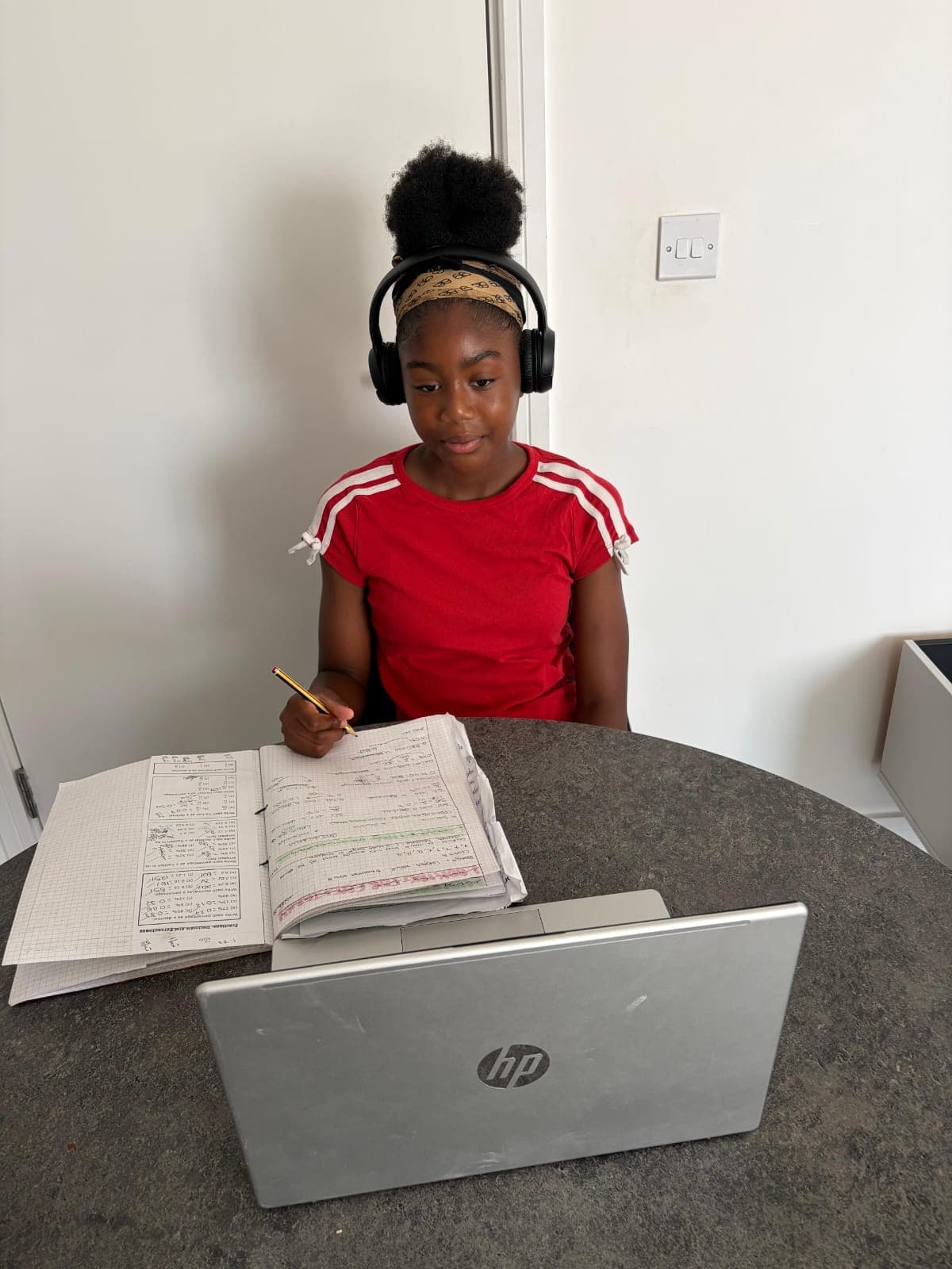 A student wearing headphones, taking notes in a notebook next to an open laptop at home